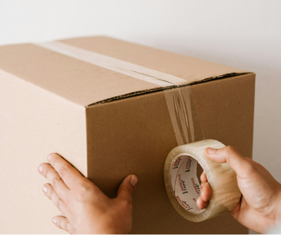 A closed cardboard box, packing tape is in the middle of being applied to seal it.